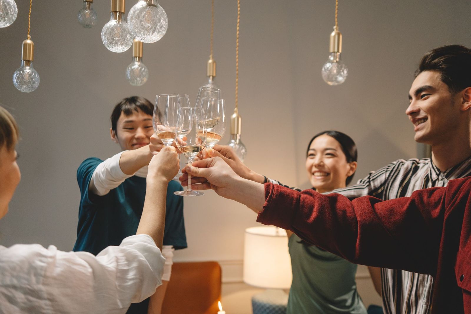 Four friends standing around a table indoors clinking wine glasses under a cluster of hanging pendant bulbs