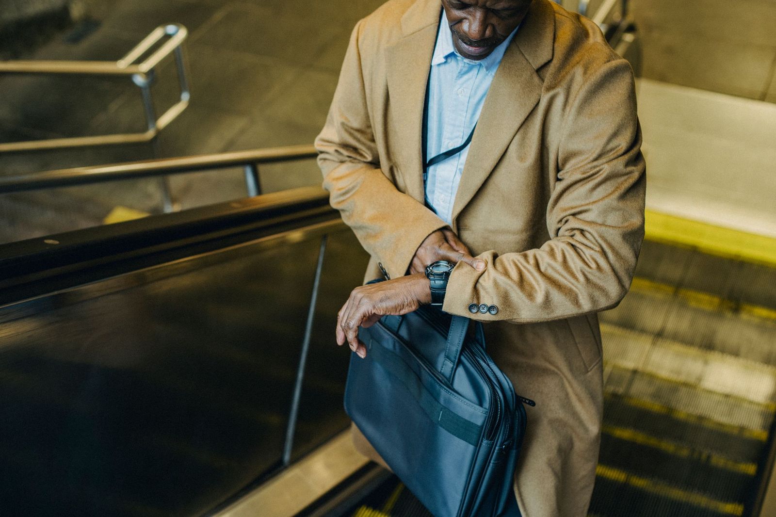 A man in a camel overcoat riding an escalator down into a station, tilting his wrist to check his watch, a laptop bag hanging from his arm