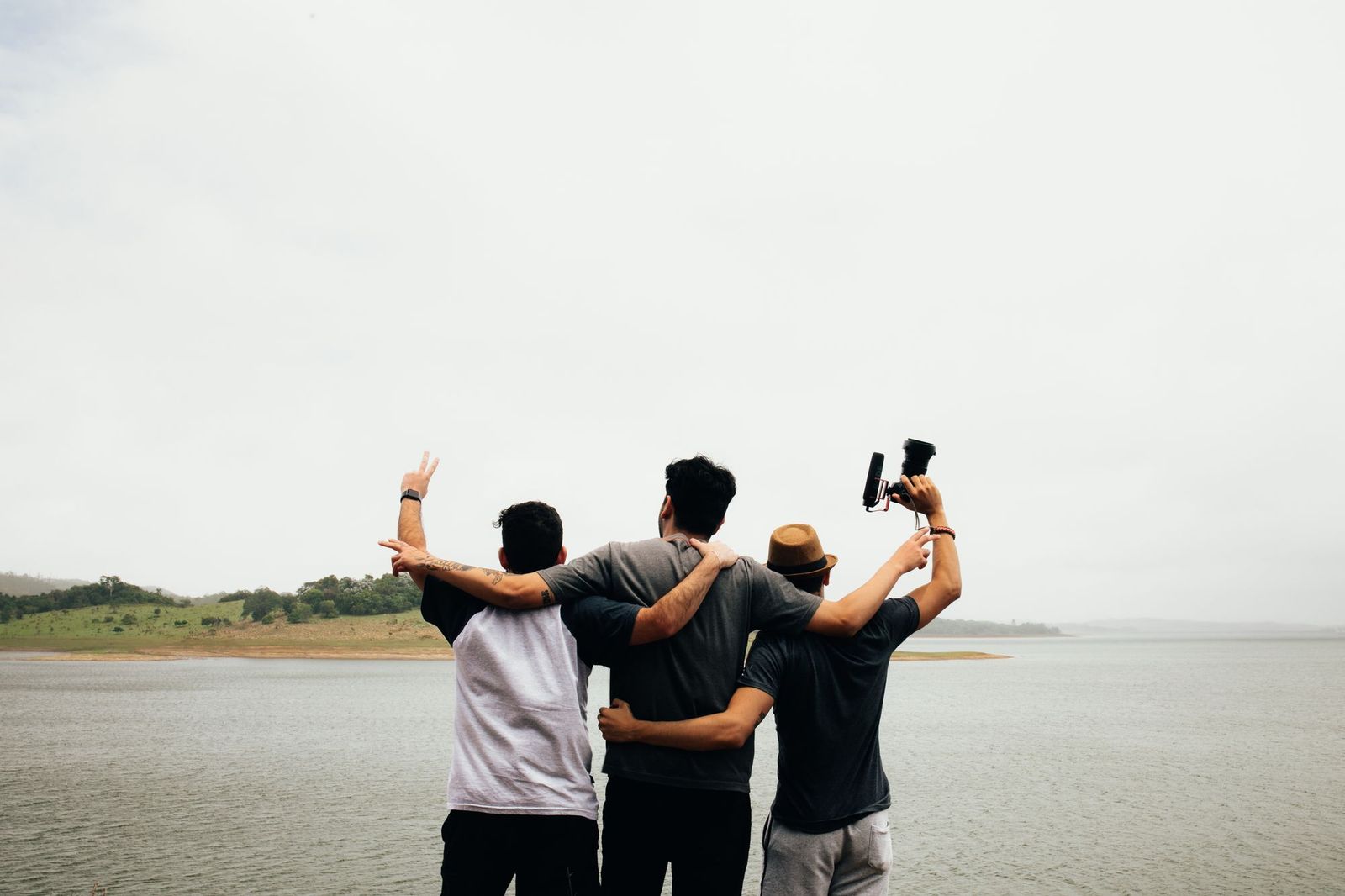 Three friends standing with their arms around each other's shoulders, seen from behind, looking out over a lake with one of them holding a camera in the air