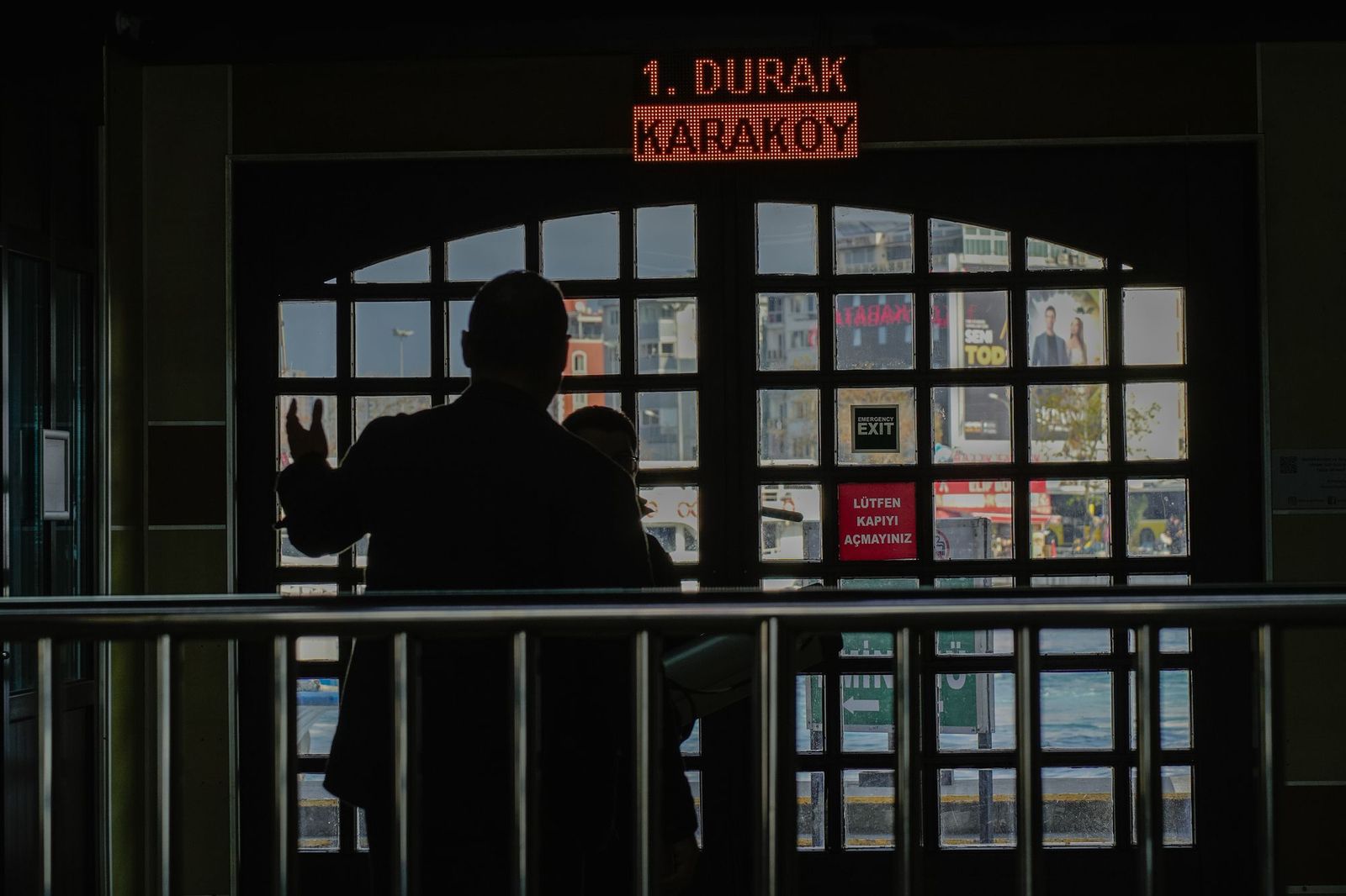 A lone figure silhouetted at a dark station entrance, one hand raised towards the glass doors, a lit sign reading Karakoy above them