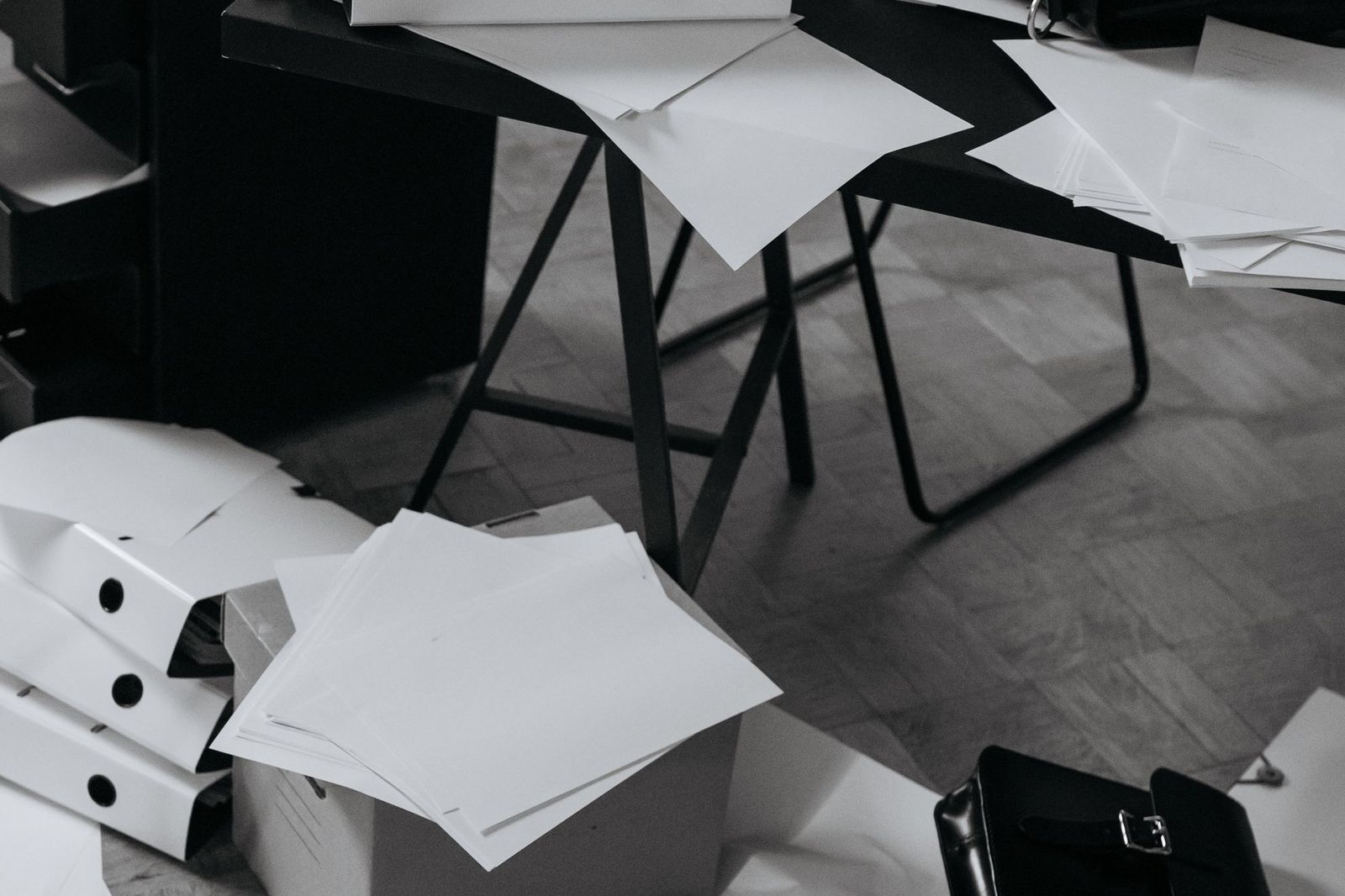 A black and white photograph of loose sheets of paper scattered across a trestle desk and the floor beneath it, next to stacked box files and a briefcase