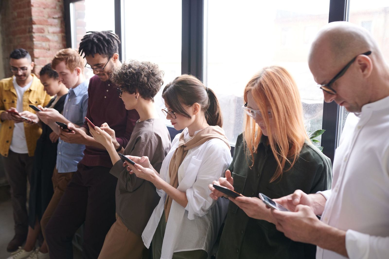 A line of people standing by a window, each looking down at their own phone and texting