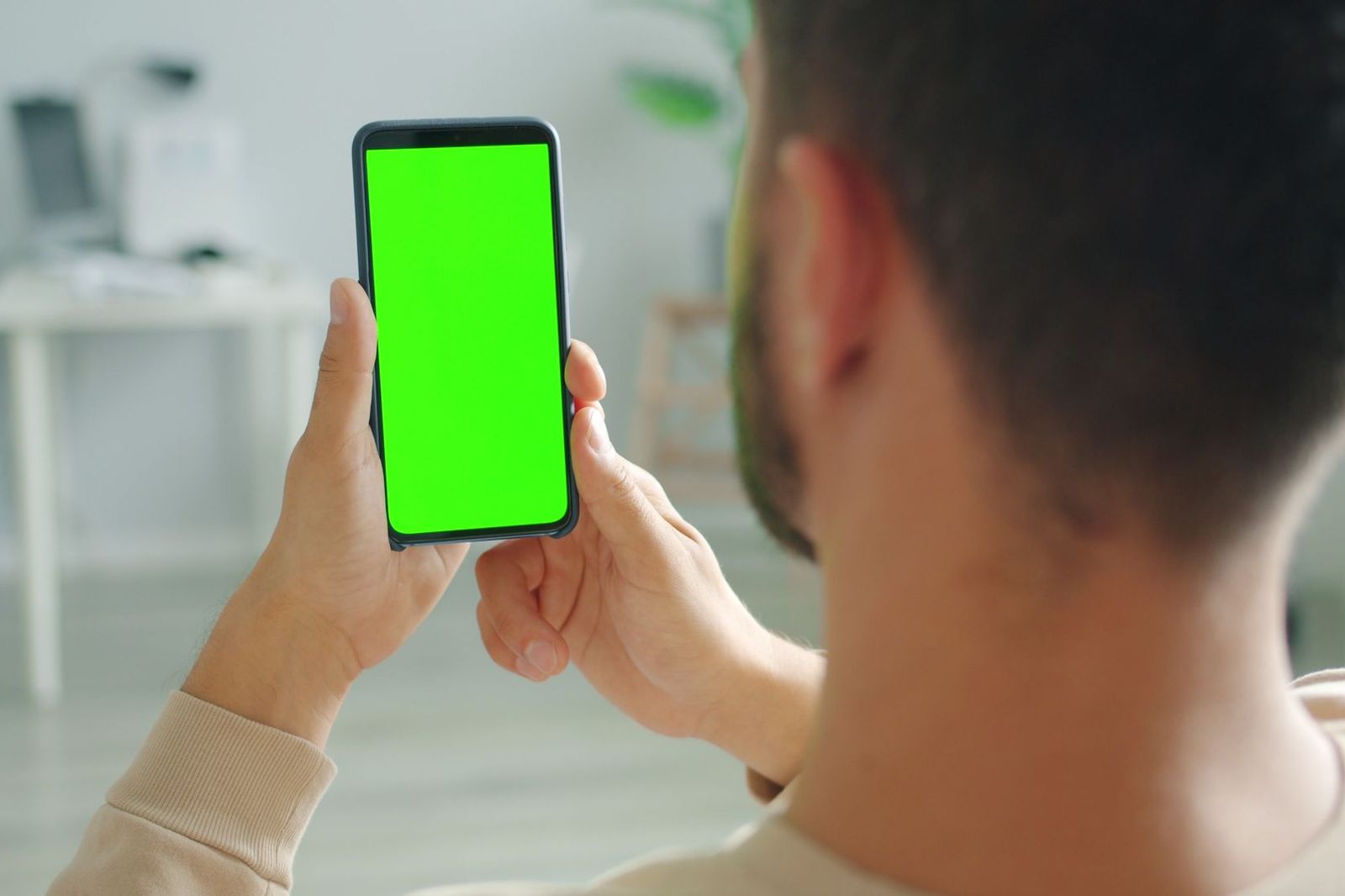 A person's hand holding a smartphone displaying a plain bright green screen, seen from over their shoulder