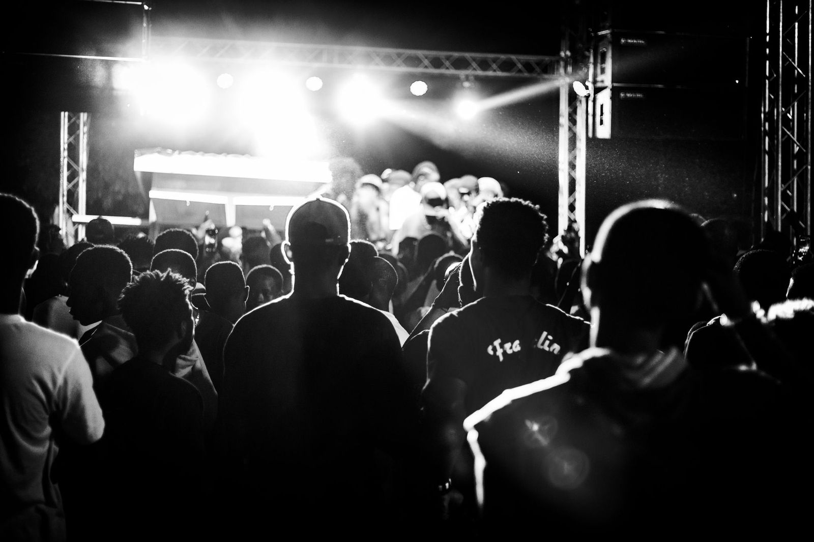 A dense crowd photographed from behind at a night concert, faces invisible, everyone silhouetted against bright stage lights
