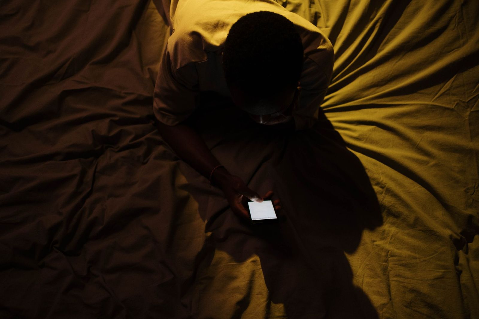 A person lying on a bed in a darkened room, face lit only by the screen of the phone they are holding