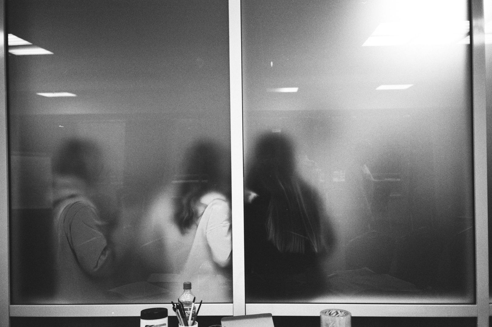 Black and white photograph of several blurred figures seen through a large pane of frosted office glass, their outlines recognisable but their faces indistinct