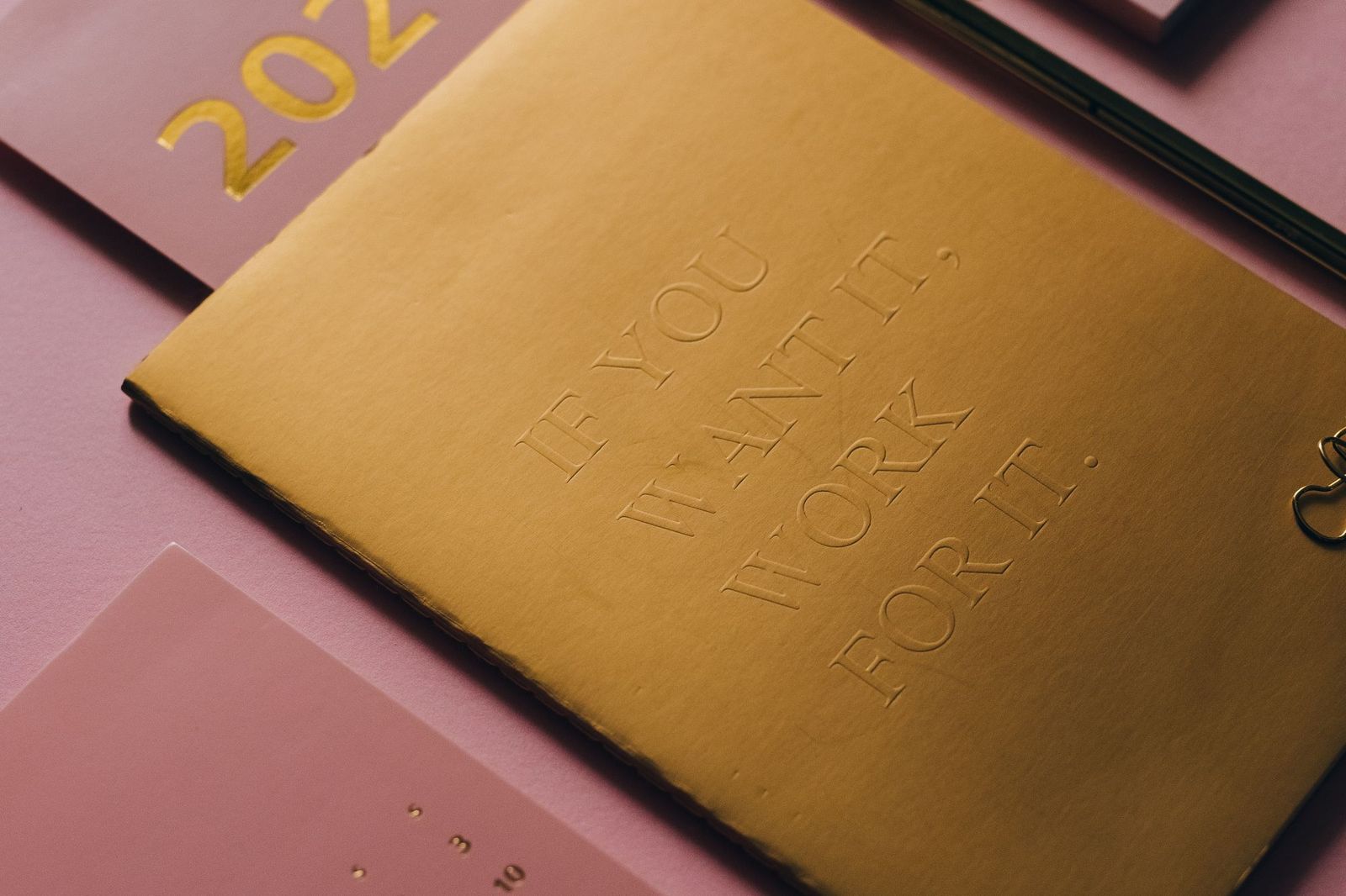 A gold debossed diary cover reading if you want it, work for it, lying on a pink desk beside a planner marked 202 in gold foil