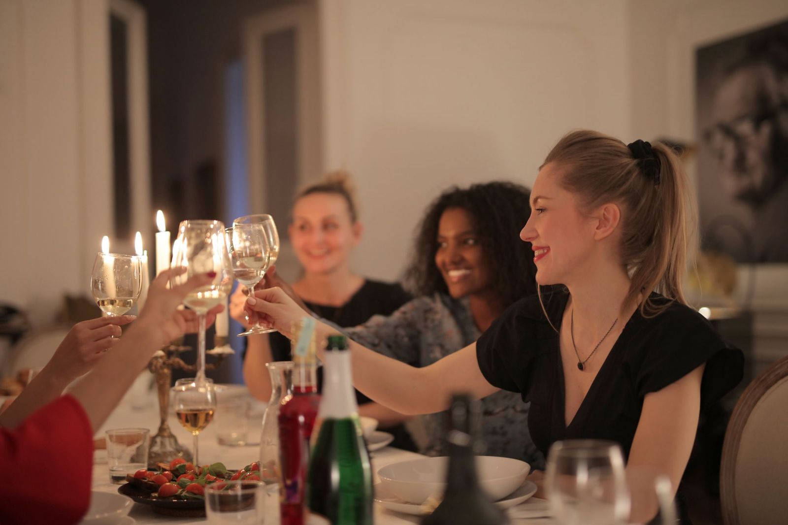 Four friends at a candlelit dinner table clinking glasses of white wine, with plates, a green bottle and a bowl of tomatoes in front of them