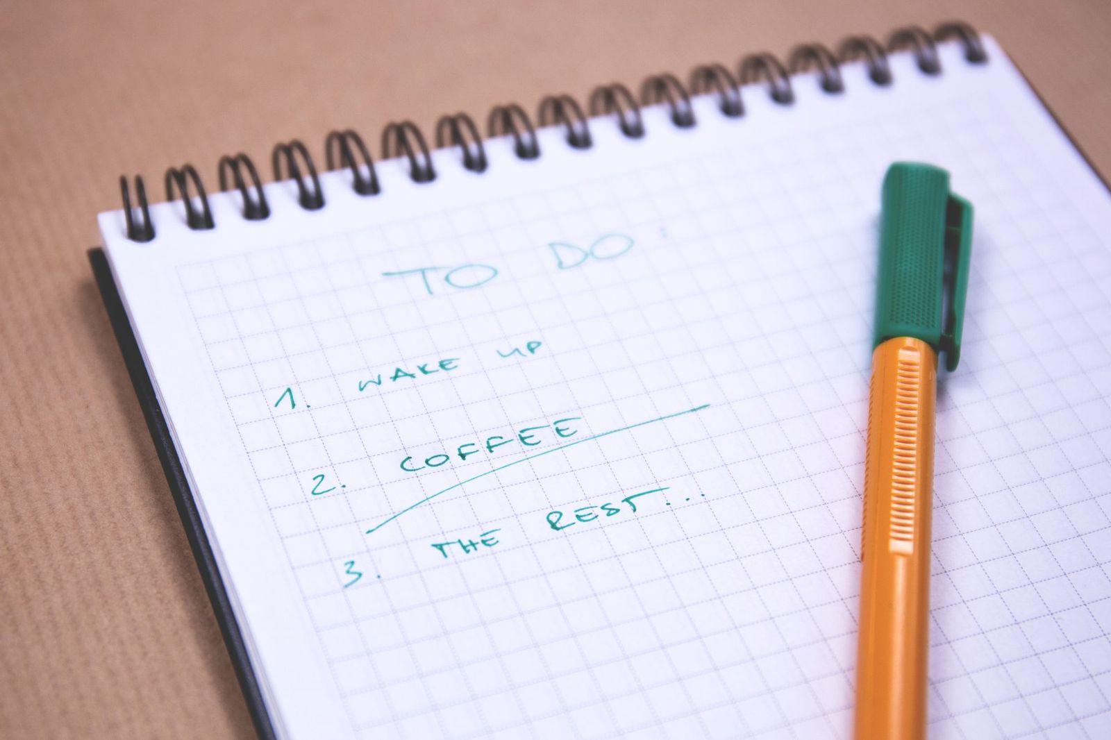 A spiral-bound squared notepad on a brown desk with a handwritten to-do list reading wake up, coffee, the rest, next to an orange pen with a green cap