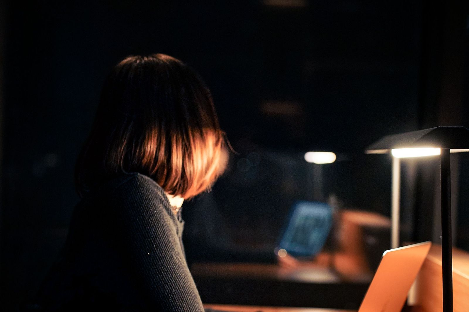 A woman sitting alone in a dark room lit only by a small lamp, head lowered towards a phone, with another person blurred in the background looking at a glowing screen